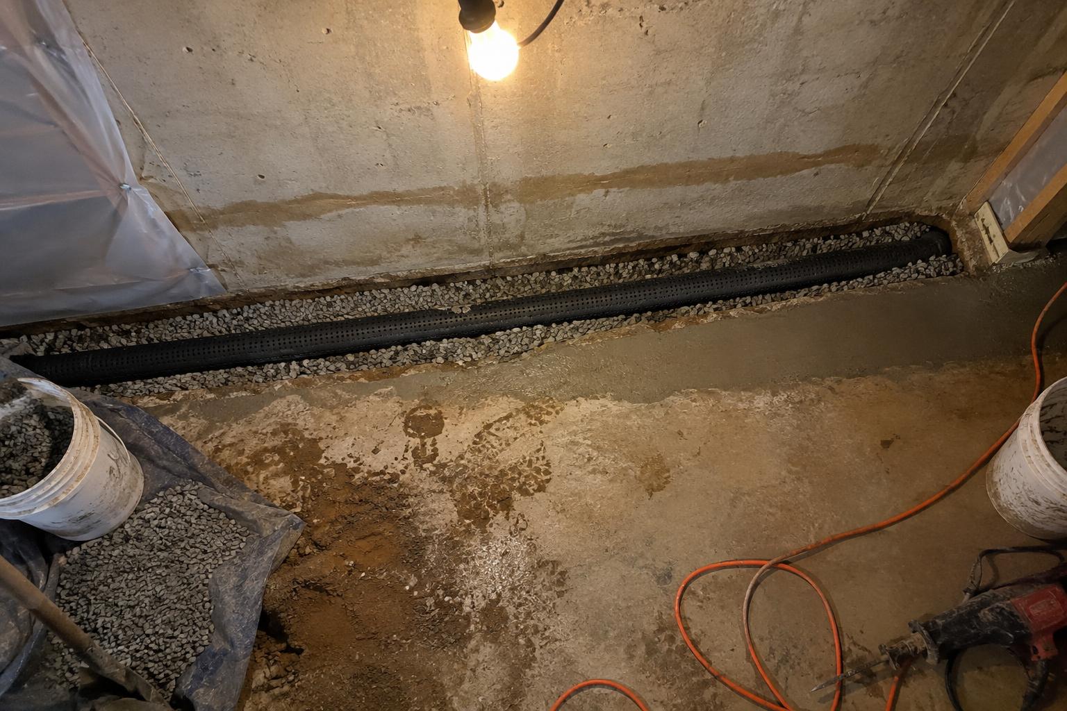 A perforated drain pipe set in clean gravel inside a trench cut along the base of a poured concrete basement wall, with a water stain line visible above it.
